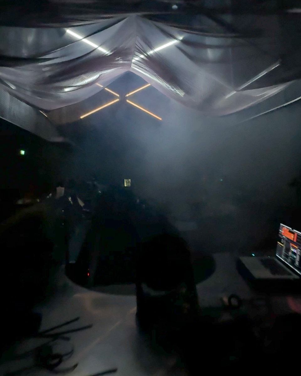 The view from behind a DJ booth, with a laptop running control software under draped fabric and haze.