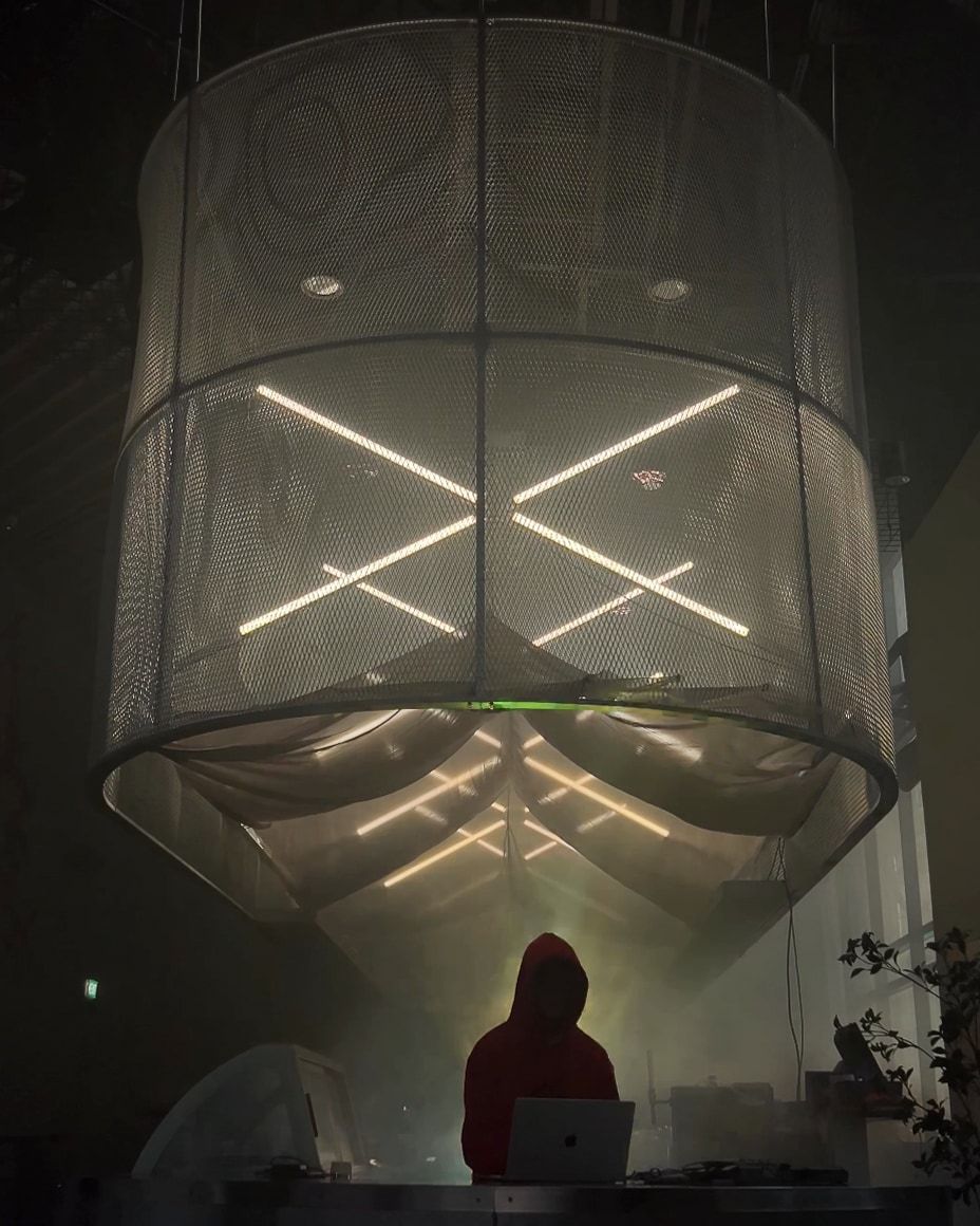 LED tubes glowing in an X shape inside a mesh drum that hangs above a DJ booth.