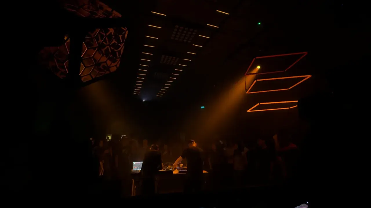 Square LED panels glowing orange along a dark ceiling above a crowd, with a red visual on a screen.
