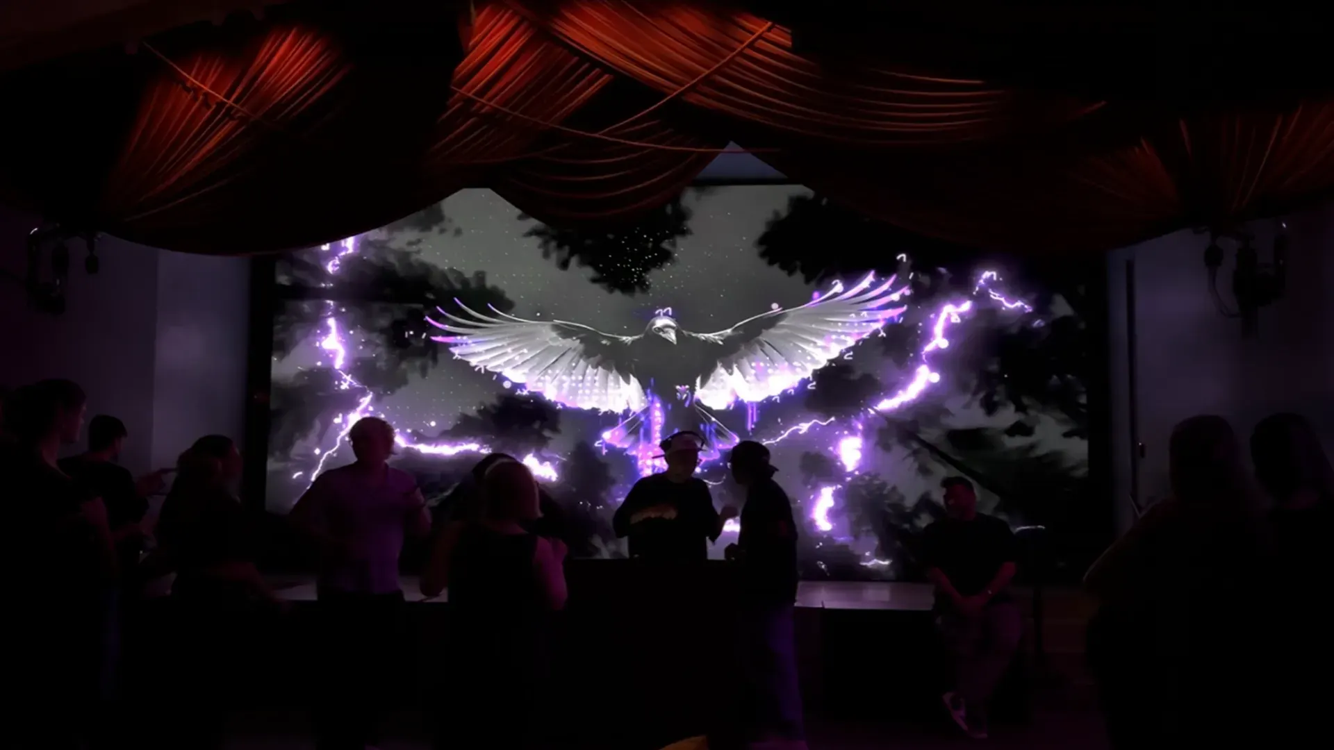 A stage under red drapes with an LED wall showing a bird and purple lightning behind the DJ.